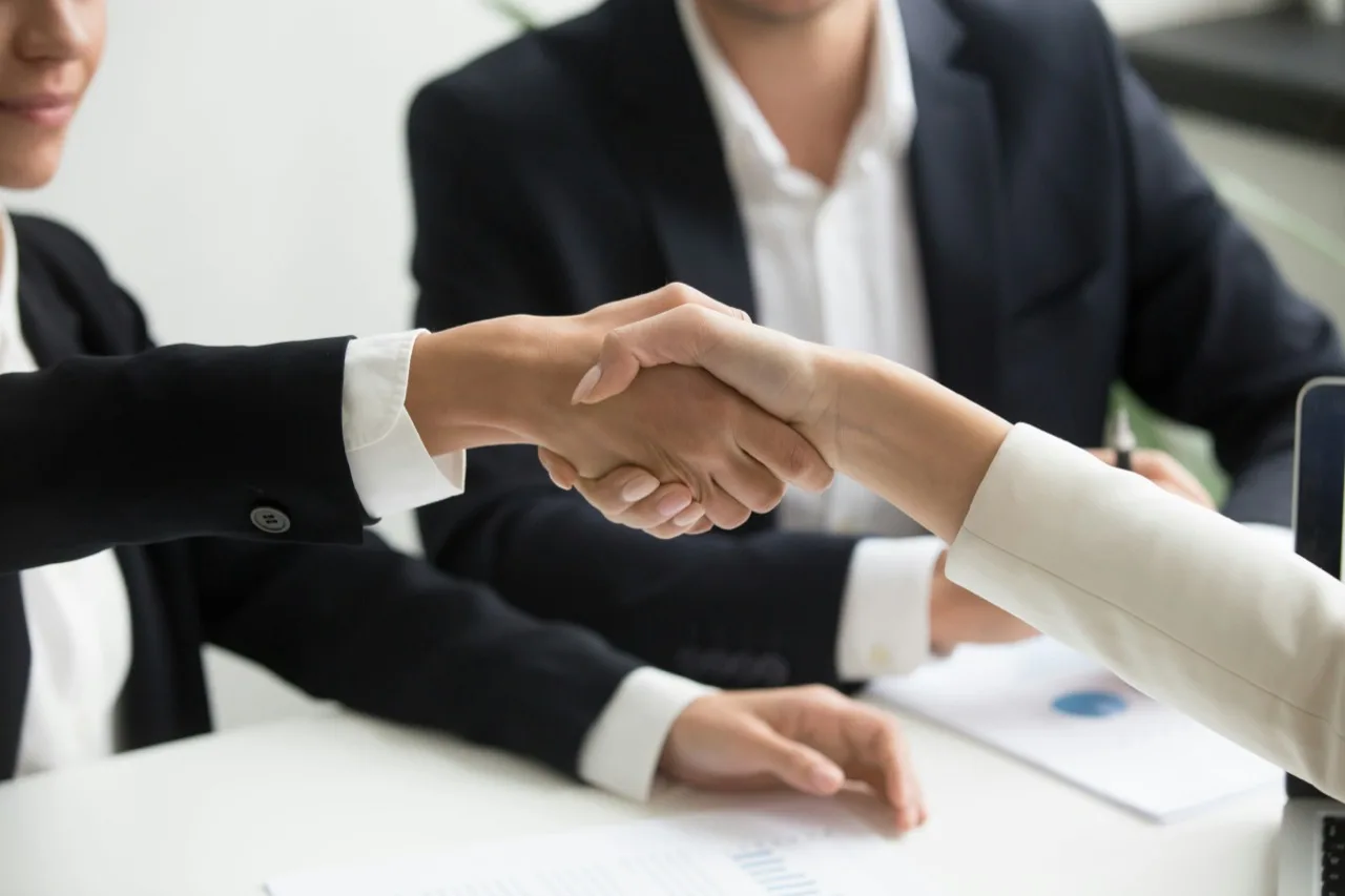 Two people in business attire shaking hands across a desk with documents on it