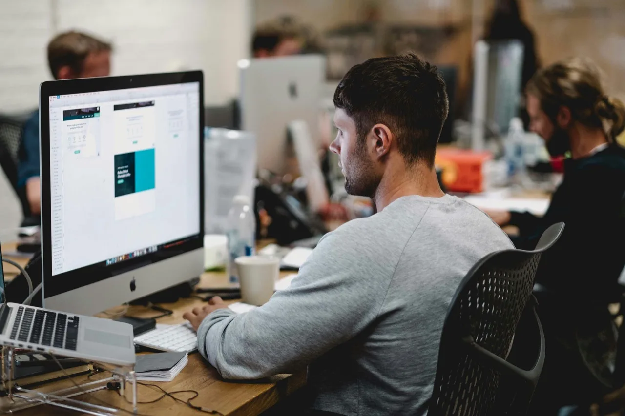 Person working at a desktop computer in an open office, teammates at desks in the background